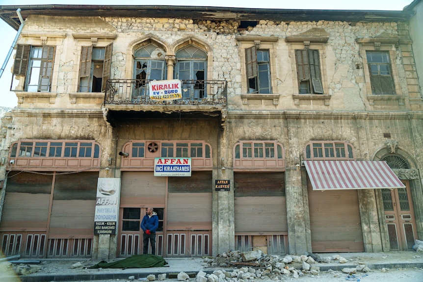 A man stands next to a tarp-covered body in front of a damaged building