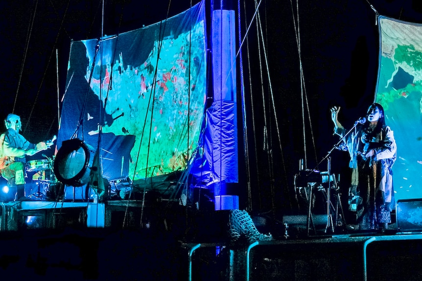 Two people performing on a sailboat at night with projections on the sails.