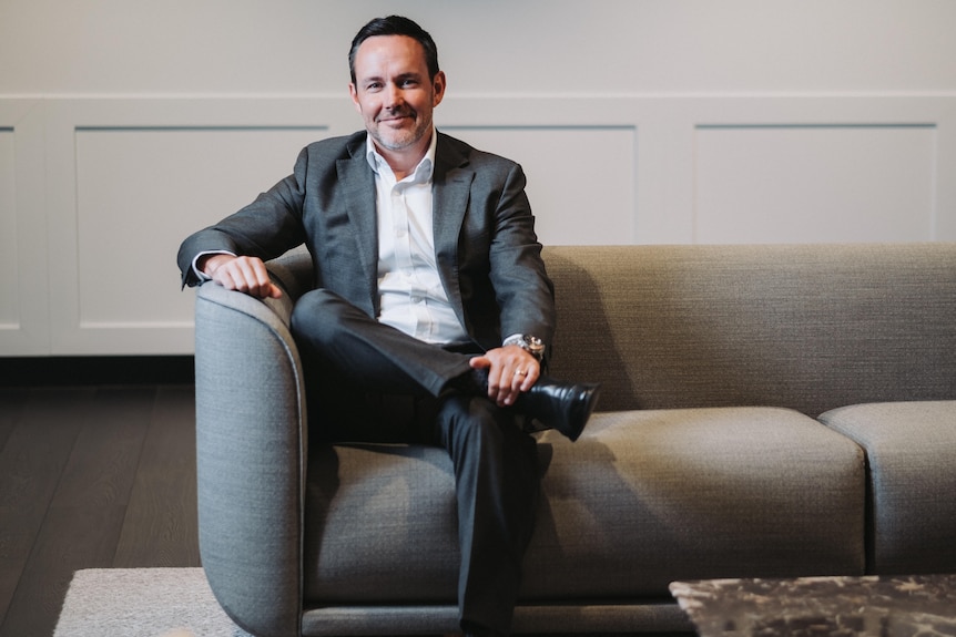 A smiling man in a grey suit, white shirt sits in a lounge in front of a white dado wall.