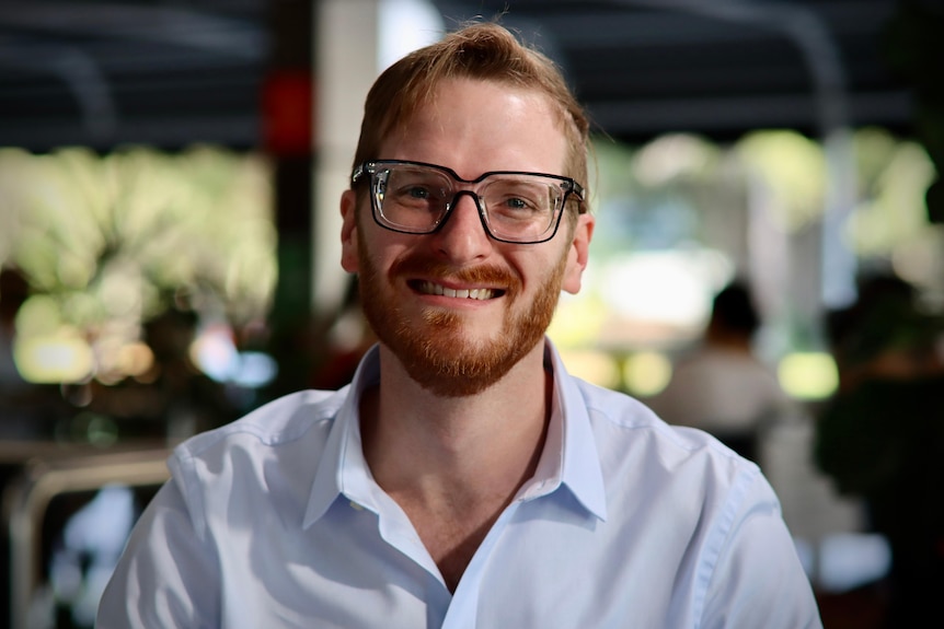 A man wearing glasses and a dress shirt smiles into the camera. 