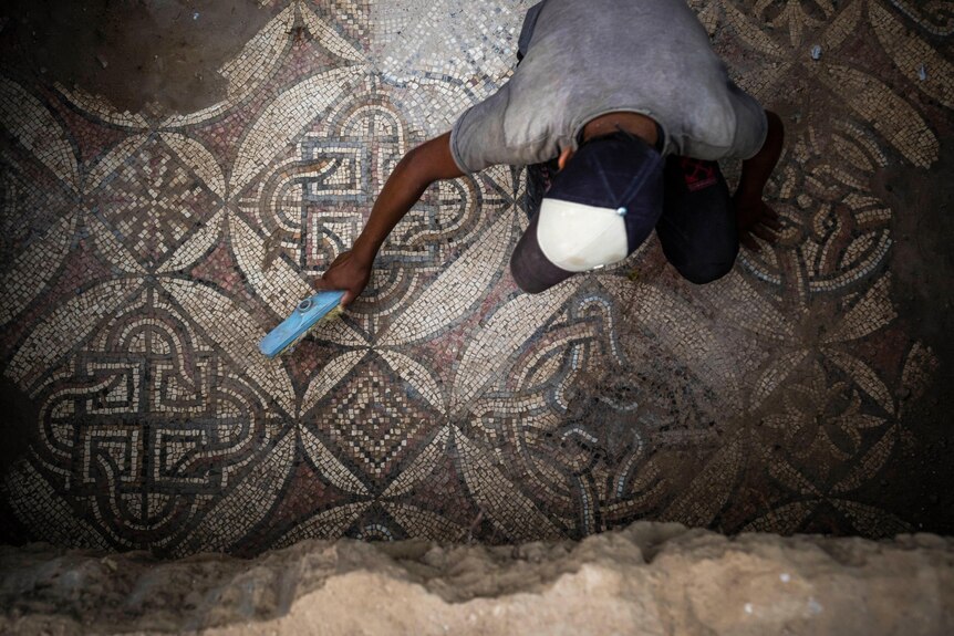 A top-down view shows a young man sweeping dust off a complex, ancient mosaic with a small brush.
