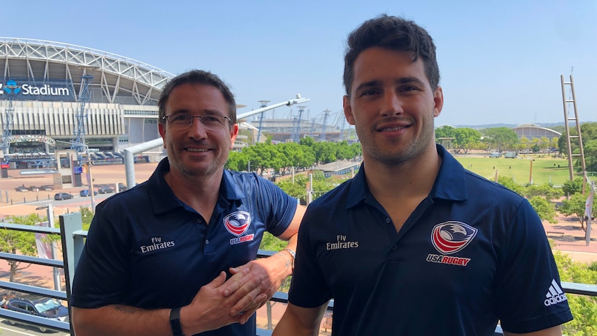 Two men stand wearing dark navy blue polo shirts with a stadium in the background.