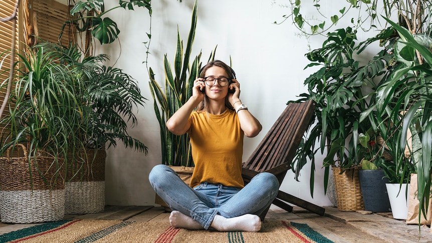 Young woman sits cross-legged in a plant-filled apartment and listens to music with a serene expression.