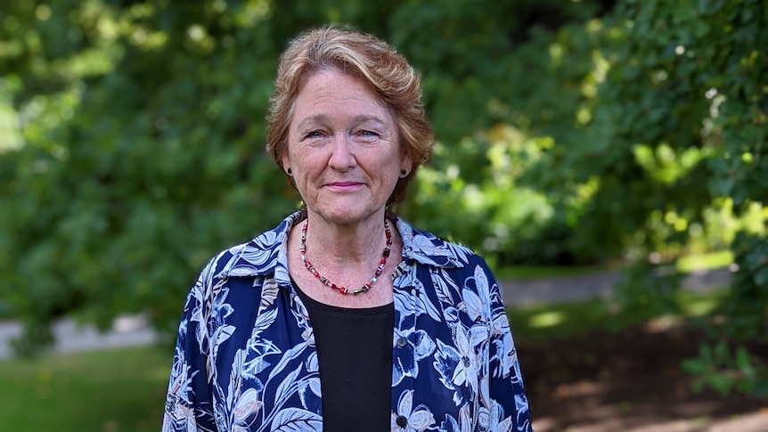 A woman wearing a colourful necklace and blue shirt, with trees in the background.