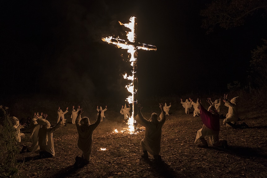 Colour still image of Ku Klux Klan members gathered in a circle around a large cross on fire at night time.