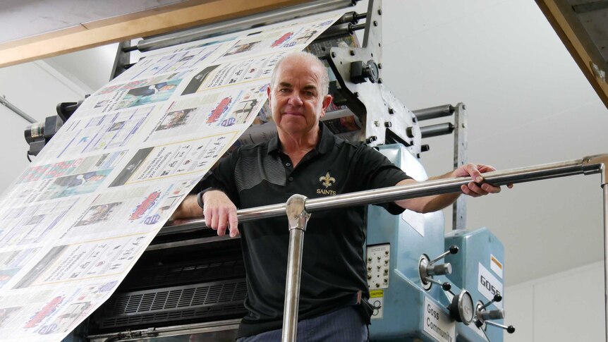 A man in a polo shirt stands next to a printing press loaded with newsprint on a mezzanine level, looking down at the camera.