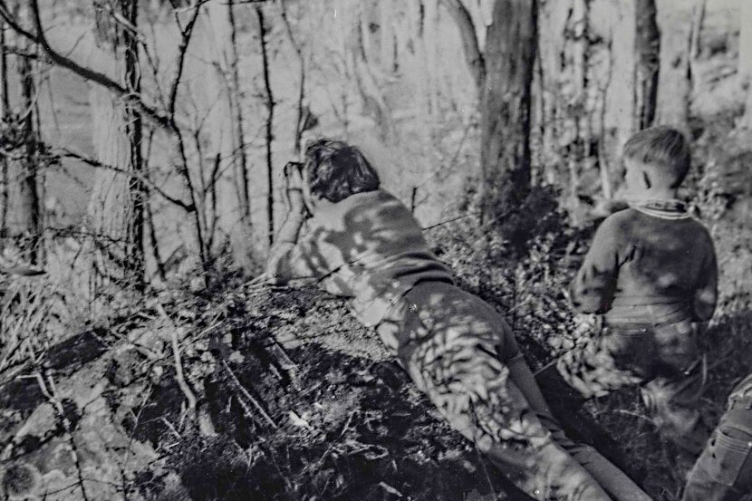 Black and white photo of a woman lying on the ground with a camera in the bush.