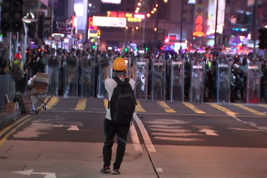 A lone protester wearing a yellow helmet faces a row of police officers holding shields.