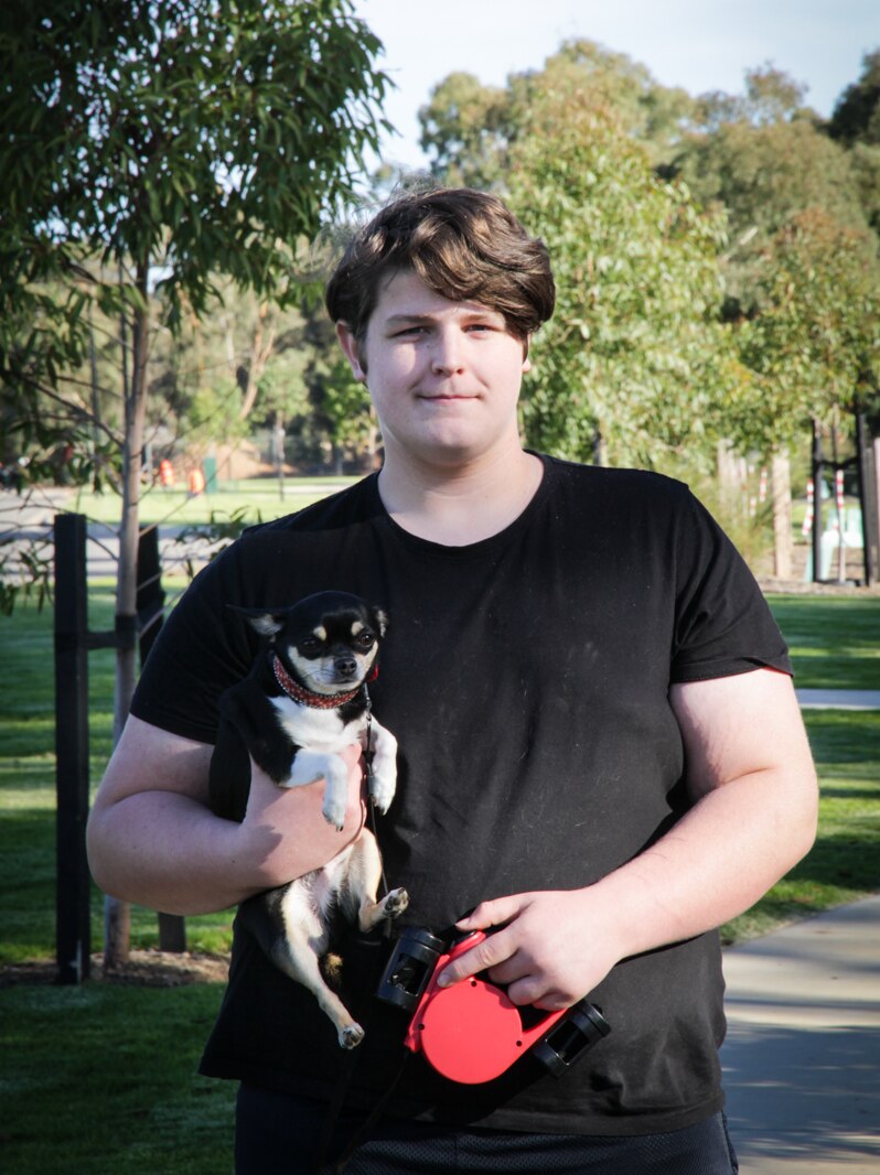 A young man wearing a black t-shirt stands smiling holding a small black and white dog.