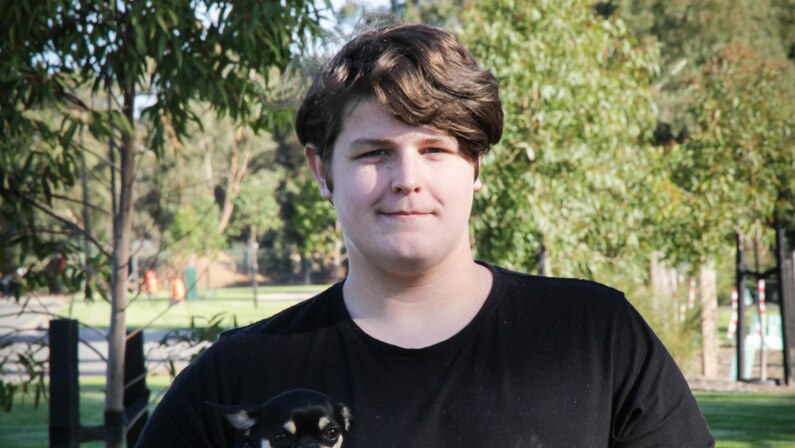 A young man wearing a black t-shirt stands smiling holding a small black and white dog.