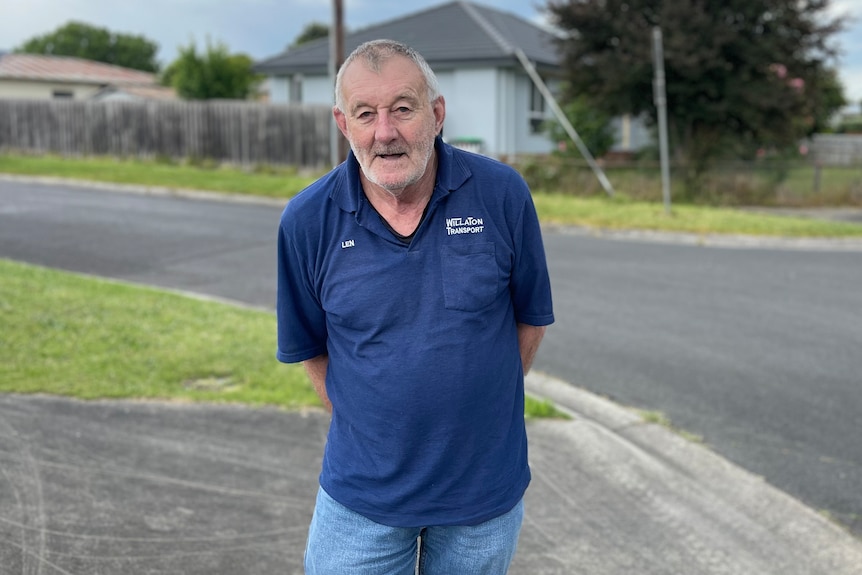 Middle aged man with grey hair in a blue shirt standing in front of an old cement sheeting house on a residential street.