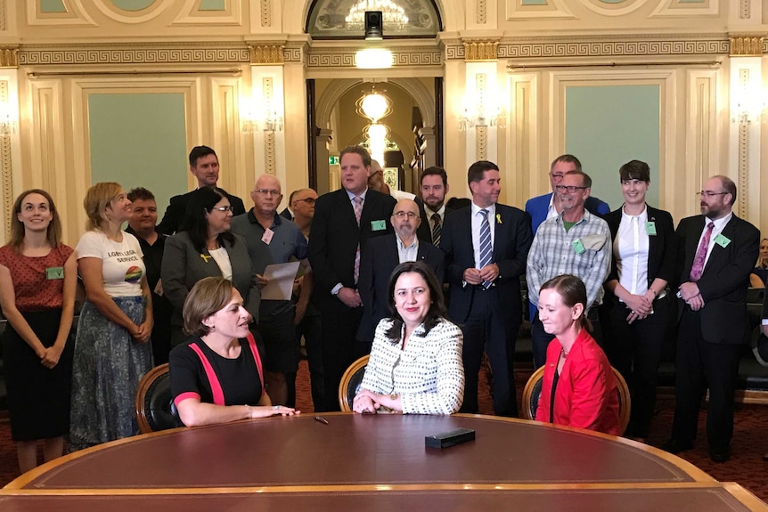 Woman in a white pattern jacket sitting in a room in parliament with a row of people behind her.