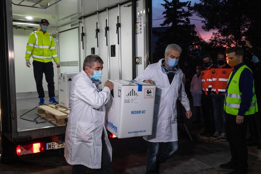 Two-middle aged men in white coats and mask carrying boxes off a truck at night as guards look on.