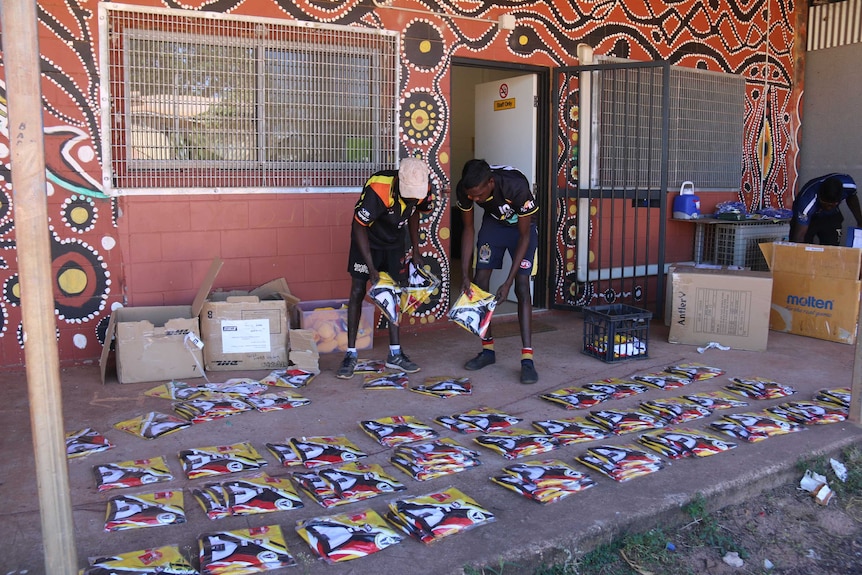Two Aboriginal men putting football jerseys into piles on the ground in front of a brick wall covered with Indigenous art.