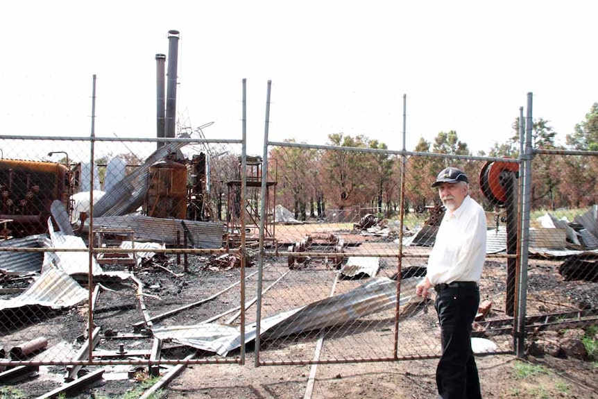 Man stands outside fencing gates looking at wreckage and twisted metal and burned engines