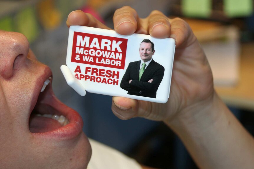 A woman holds to her mouth a container of mints marked Mark McGowan and WA Labor A Fresh Approach.