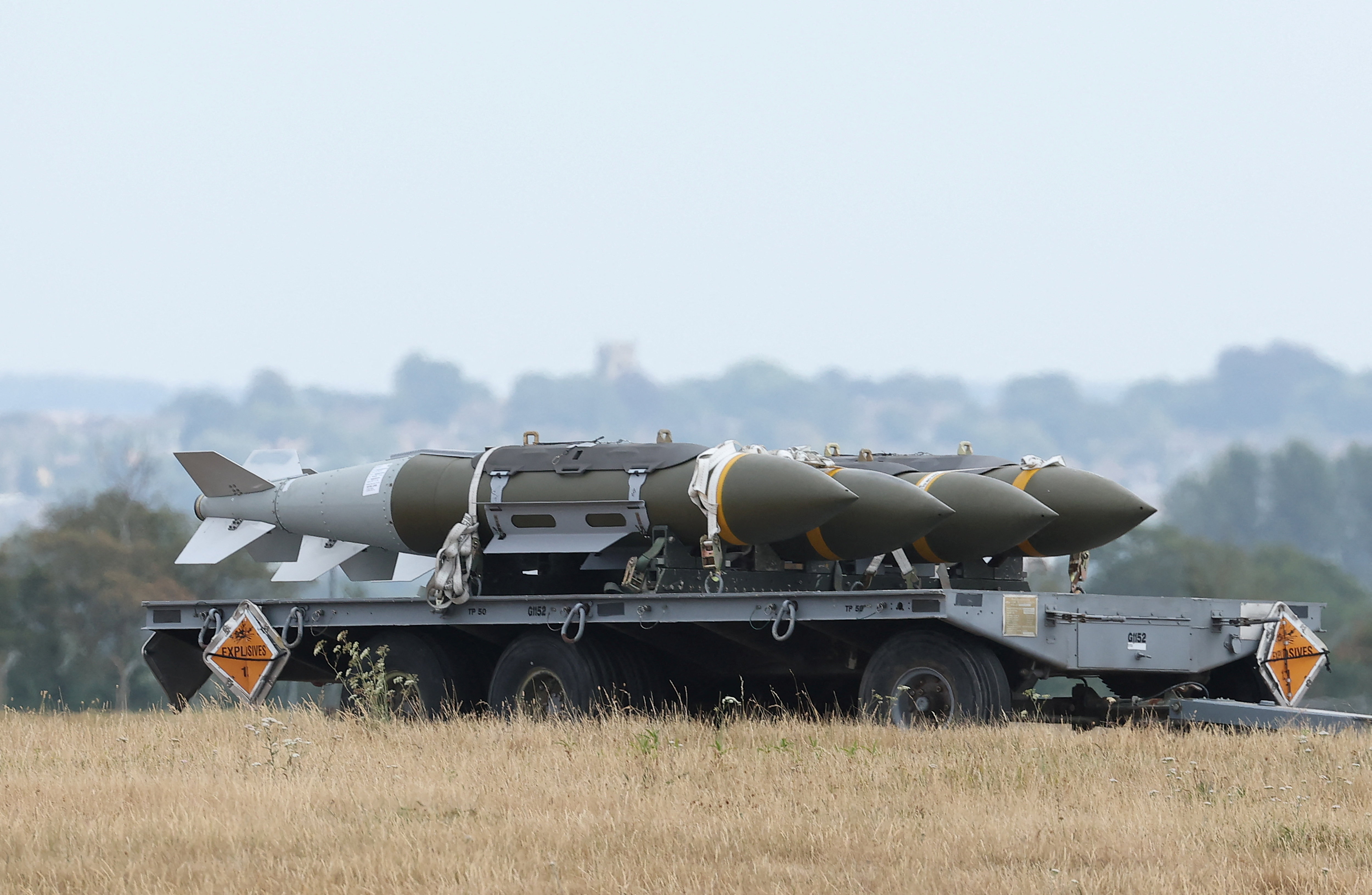 Three large, olive green rocket munitions sit on the back of a grey trailer trolley in an open field.