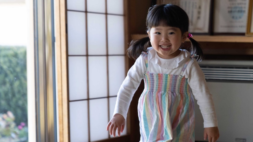 A little Japanese girl in pigtails and a rainbow coloured dress runs through a room