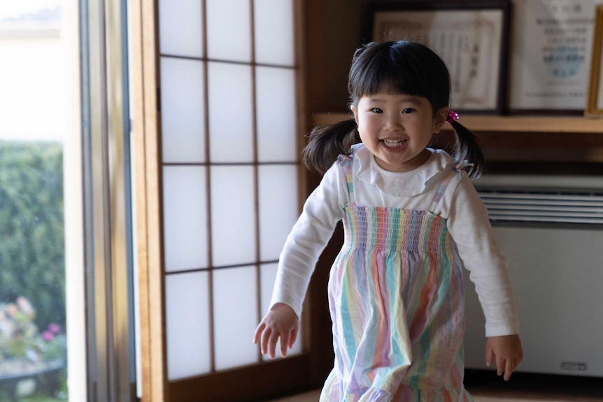 A little Japanese girl in pigtails and a rainbow coloured dress runs through a room