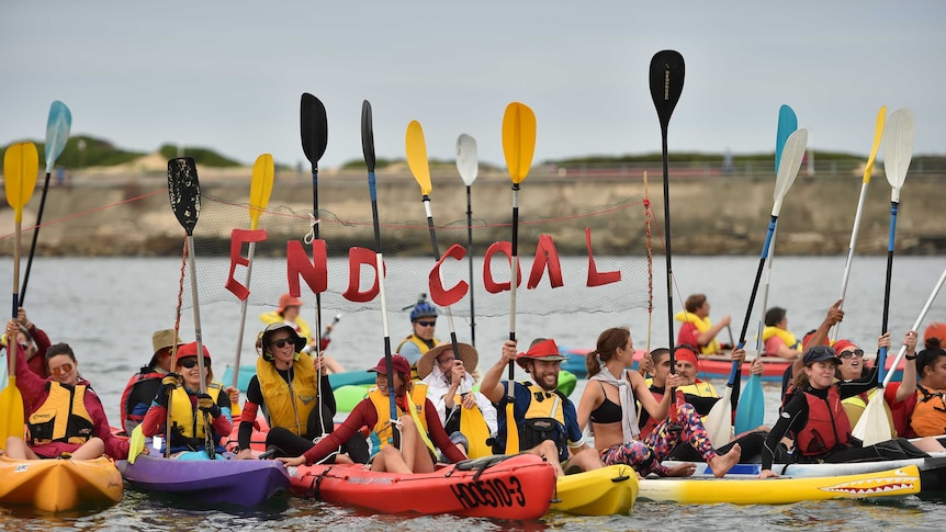 A group of people in kayaks float on a harbour holding a sign that says 'no coal'.
