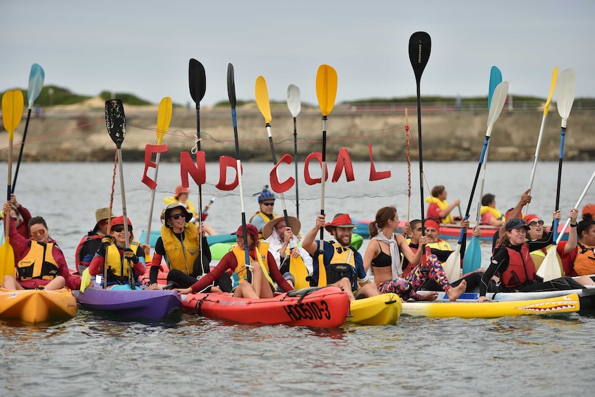 A group of people in kayaks float on a harbour holding a sign that says 'no coal'.