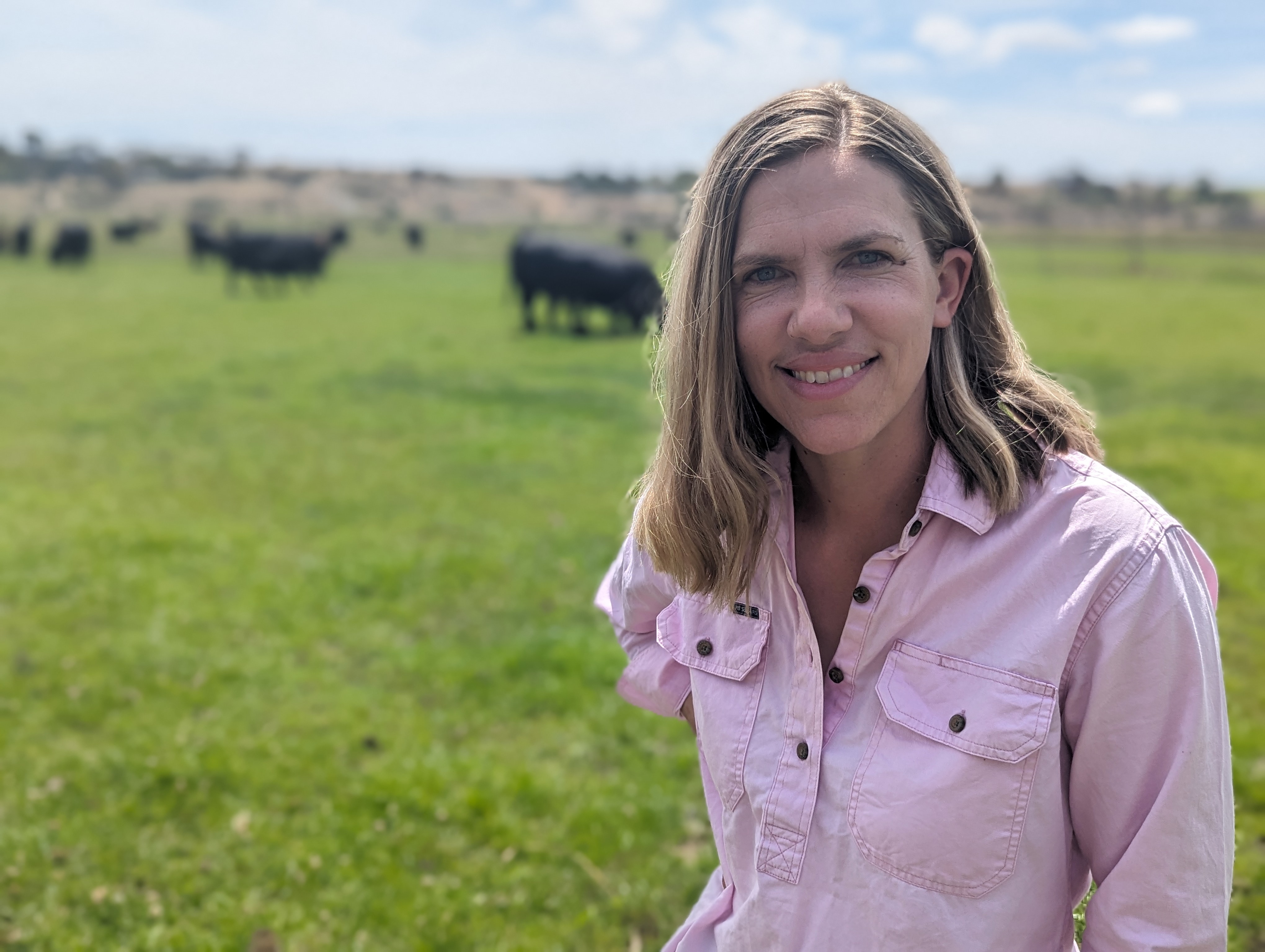 A smiling blonde woman, Alex, smiles, with beef cattle in the background on her lush paddock.