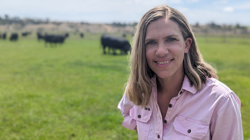 A smiling blonde woman, Alex, smiles, with beef cattle in the background on her lush paddock.