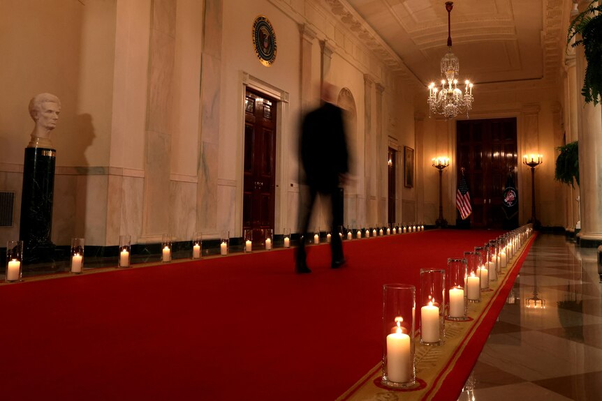 U.S. President Joe Biden (blurred by a slow shutter speed) walks down a hallway lined with candles 