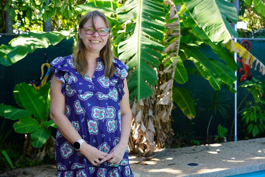 Woman wearing a patterned blue dress and red glasses smiles, with her hands together. Tropical trees are in the background.