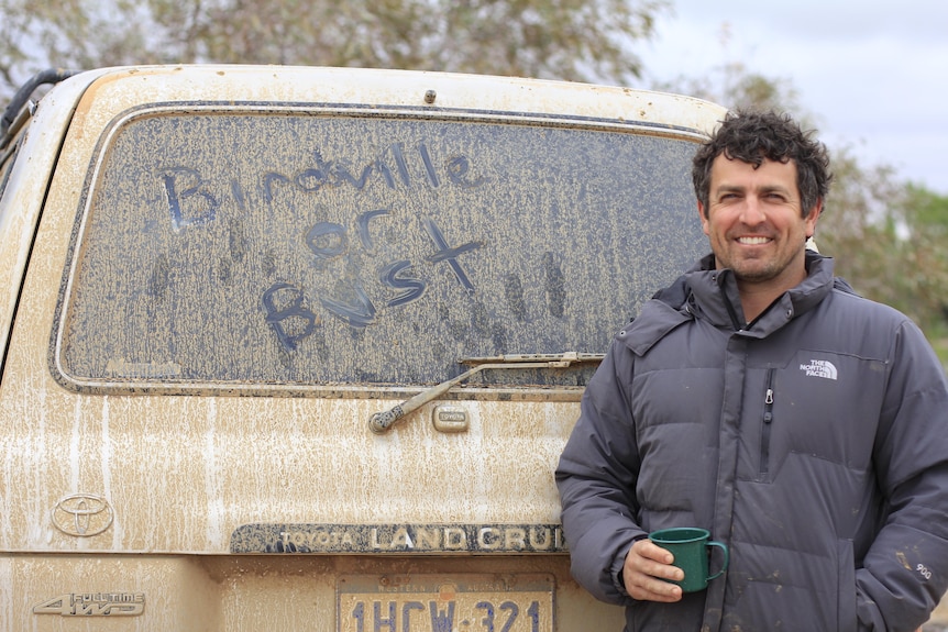 A man in a puffer jacket smiles next to his cars rear windscreen which says 'Birdsville or Bust'