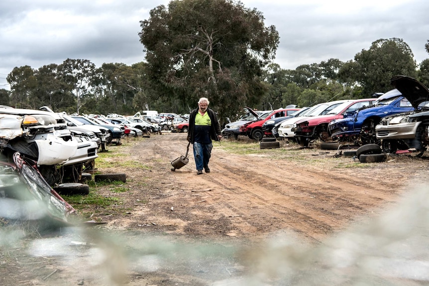 A man drags a muffler down a dirt road surrounded by wrecked cars at the auto wreckers.