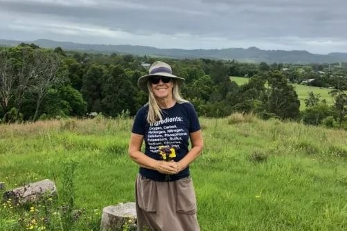 A woman wearing sunglasses and a hat hold flowers in a paddock. Green trees and hills are behind her. 