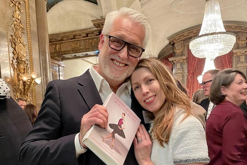 A man and woman stand side by side. The man holds a book cover towards the camera