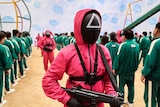 A person in a pink outfit with a triangle on their black mask holds a gun.
