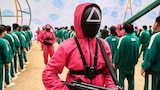 A person in a pink outfit with a triangle on their black mask holds a gun.