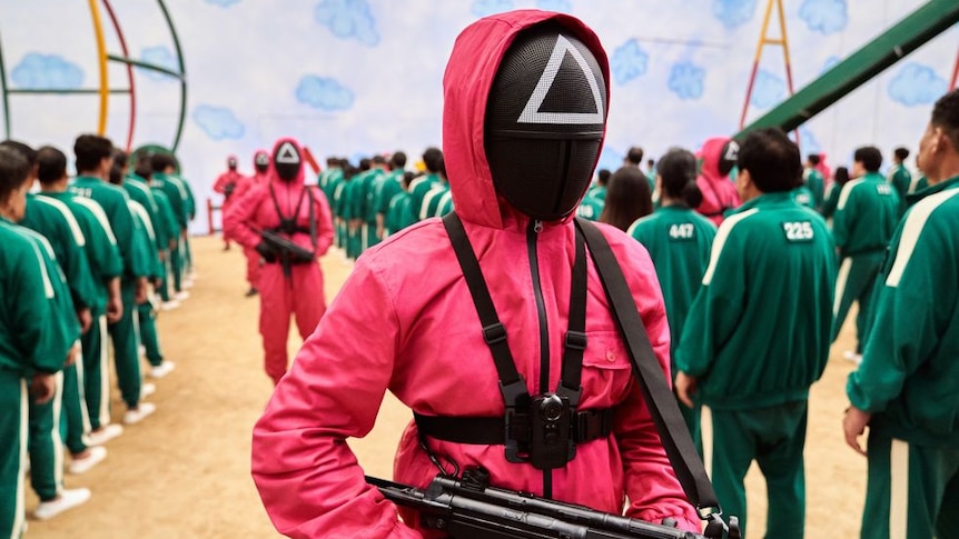 A person in a pink outfit with a triangle on their black mask is holding a gun.