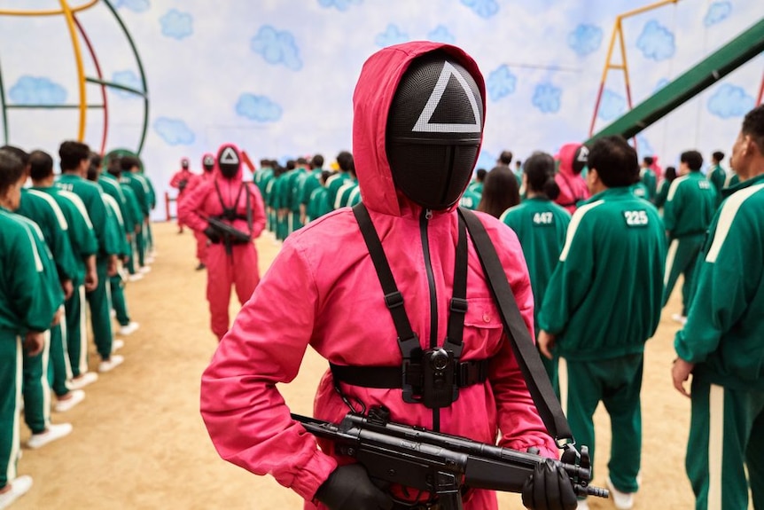 A person in a pink outfit with a triangle on their black mask holds a gun.