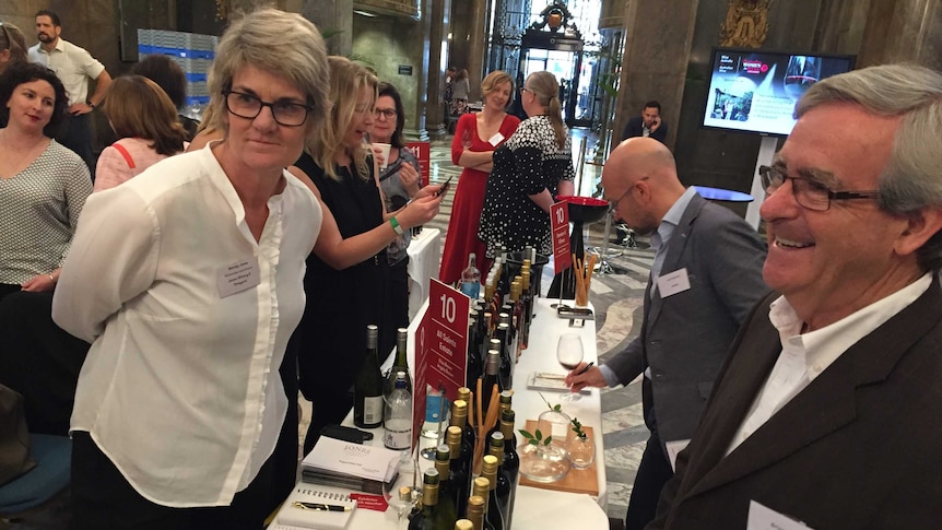 A grey-haired man laughs with multiple wines available at a long table.