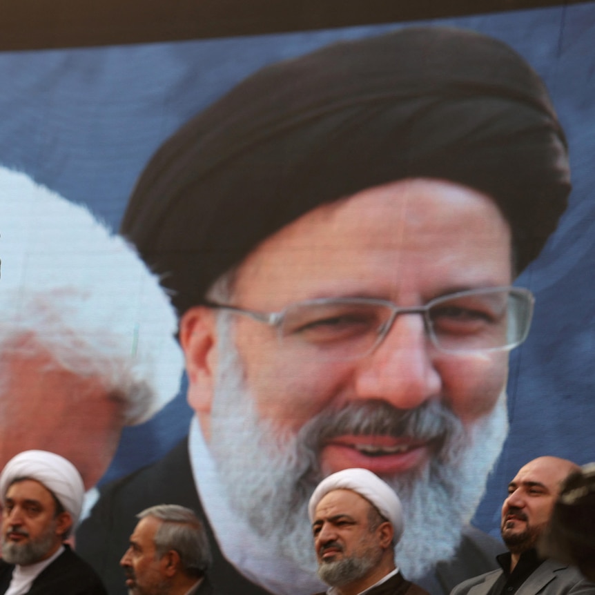 A man with a beard and glasses stands at a podium in front of a banner with the image of late Iranian president Ebrahim Raisi.