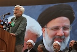A man with a beard and glasses stands at a podium in front of a banner with the image of late Iranian president Ebrahim Raisi.