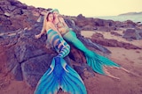 Professional mermaid performers Amelia Lassetter and Jessica Bell recline on rocks.