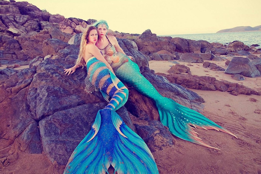Professional mermaid performers Amelia Lassetter and Jessica Bell recline on rocks.