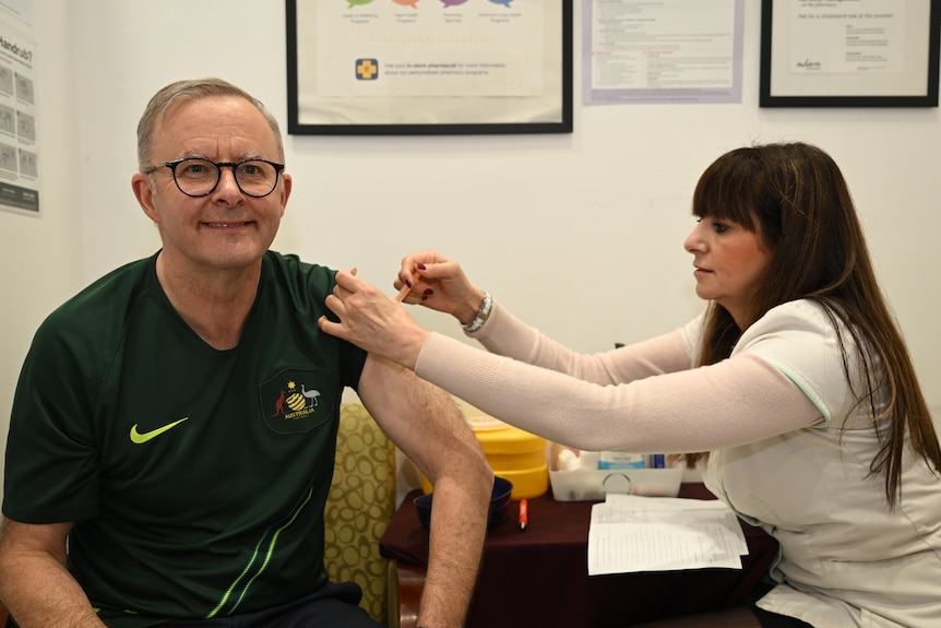 Prime Minister Anthony Abanese in an Australian football jersey receives a COVID-19 booster injection from a woman.