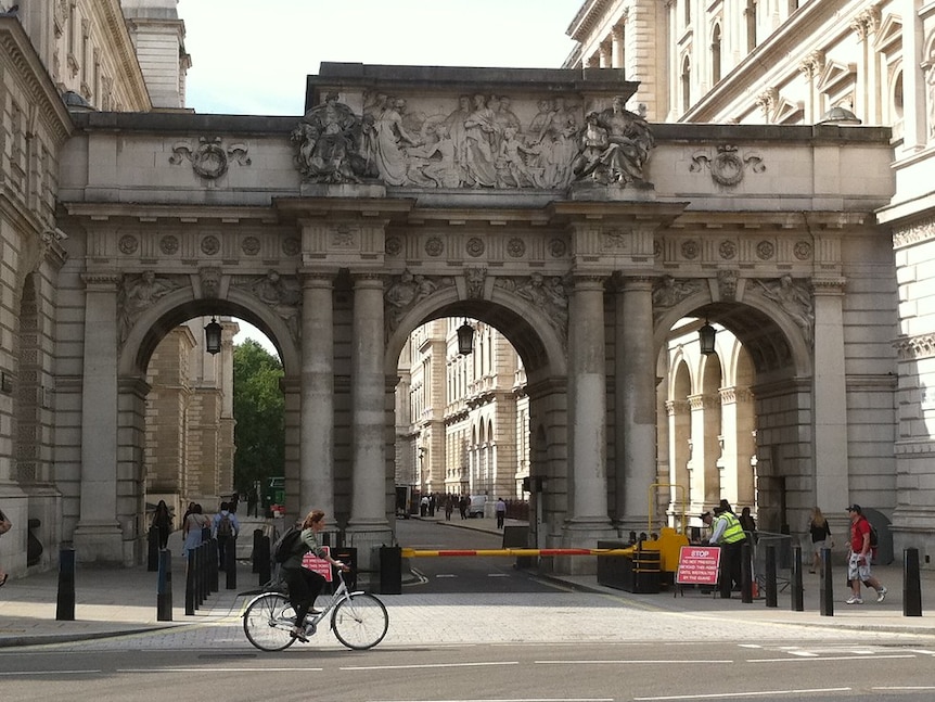 Looking across a London street, you see an ornate stone arch with figures carved at its apex as a cyclist rides in front of it.