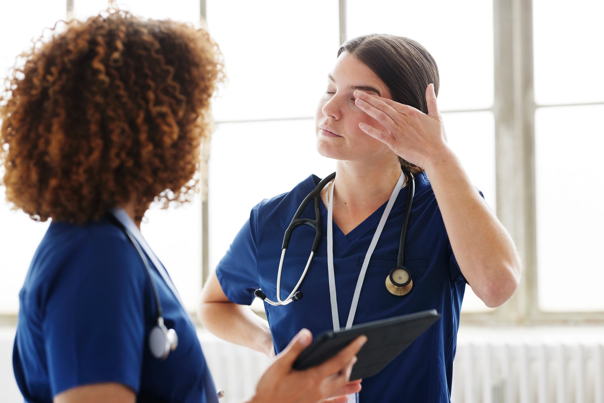 Two healthcare professionals in conversation. Both are wearing scrubs. One woman is rubbing her eyes as she's tired. 