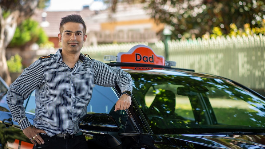 A man in a taxi driver uniform leans against his cab which has "Dad' written on the dome. 