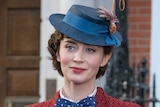 Emily Blunt in costume as Mary Poppins, wearing a red chevron-patterned coat, blue polka-dotted bow tie and shirt, and blue hat.