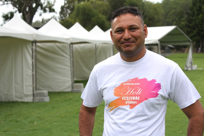 An Indian man in a white, pink and orange t-shirt standing in a park in front of white marquees.