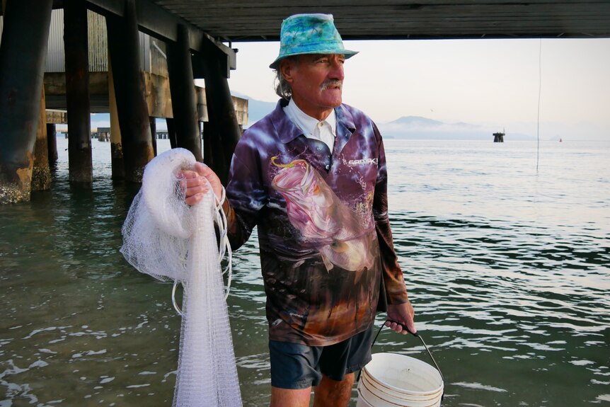 A man holding a bucket and fishing net stands in the water under a jetty