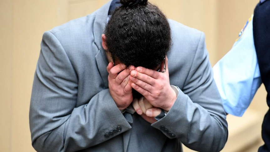 A man covers his face, as he is held by a corrections officer.